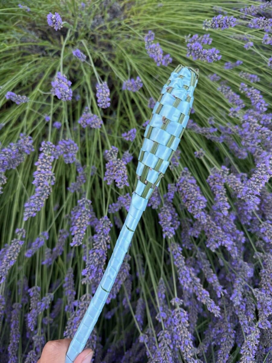 A finished ribbon-woven lavender wand held against blooming lavender at Tolt Hill Family Farm